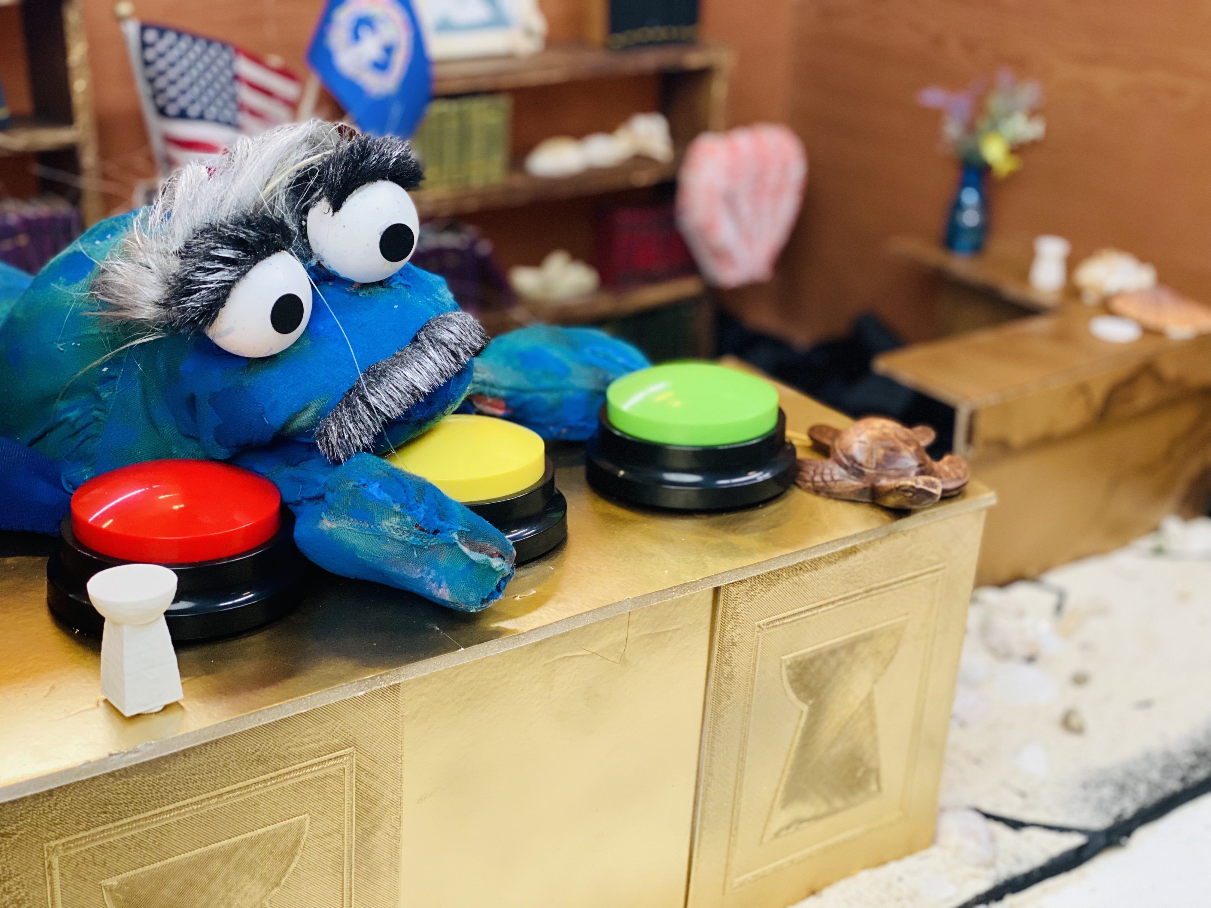 A cute, bright blue coconut crab puppet with bushy eyebrows sits at a tiny desk over three brightly colored buttons.