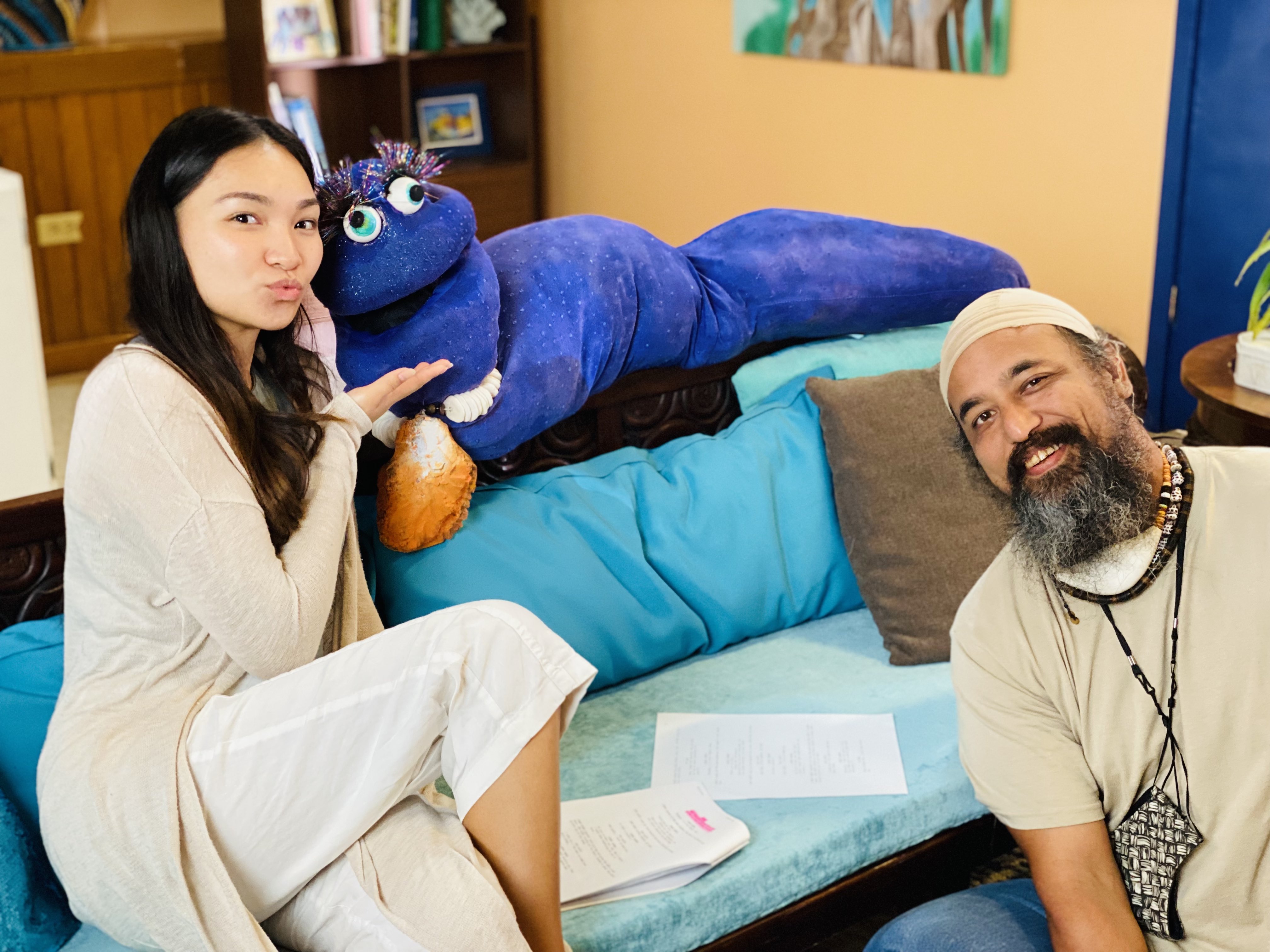A young girl with long, dark hair sits on a light blue sofa next to a large, sparkly indigo-colored dudus balati, and a bearded man in a white cap and beige shirt sitting on the floor.